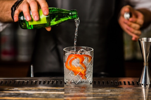 Camarero agregando gin tonic en el vaso con cubitos de hielo y piel de naranja pelada