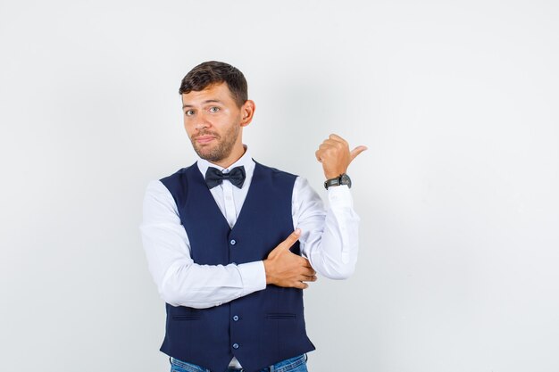 Camarero apuntando con el pulgar a un lado en camisa, chaleco, jeans y mirando confundido, vista frontal.