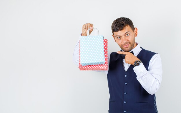 Camarero en camisa, chaleco apuntando a bolsas de papel y mirando alegre, vista frontal.