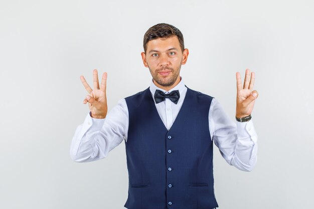 Camarero en camisa, chaleco gesticulando con tres dedos y mirando suave, vista frontal.