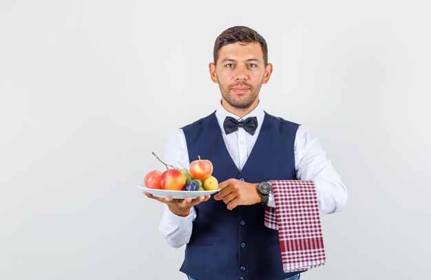 Camarero en camisa, chaleco, pajarita, sosteniendo un plato lleno de frutas y mirando alegre, vista frontal.