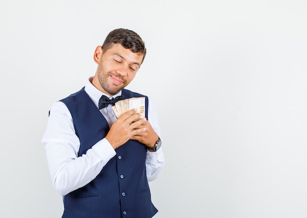 Camarero en camisa, chaleco sosteniendo dinero en el pecho y mirando esperanzado, vista frontal.