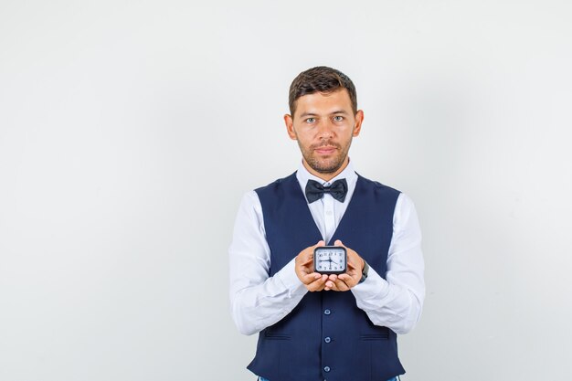 Camarero en camisa, chaleco sosteniendo reloj y sonriendo, vista frontal.