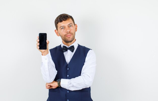 Camarero en camisa, chaleco sosteniendo teléfono móvil y mirando confiado, vista frontal.