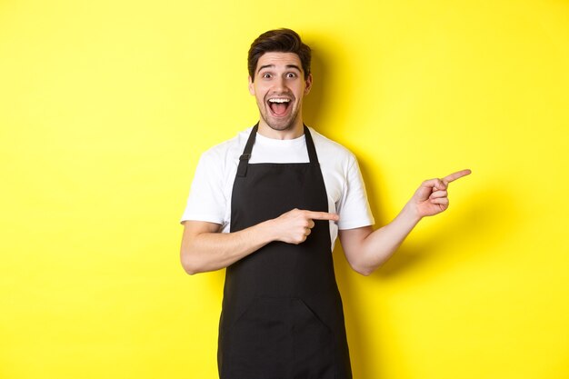 Camarero en delantal negro apuntando con el dedo hacia la derecha, mostrando publicidad y sonriendo emocionado, de pie contra el fondo amarillo