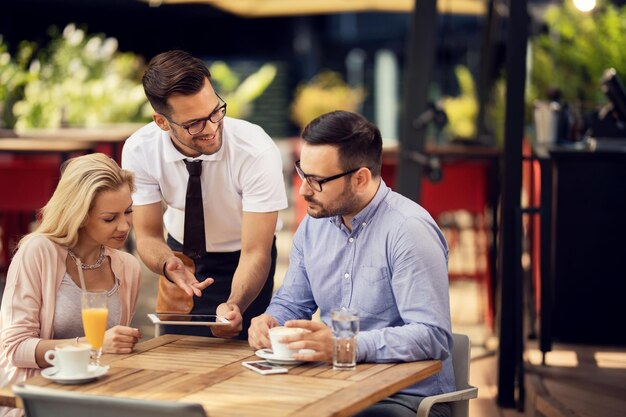 Camarero feliz hablando con los invitados mientras usa una tableta digital y mostrándoles el menú en un restaurante