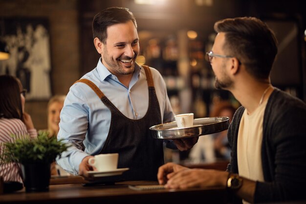 Camarero feliz sirviendo café y comunicándose con un invitado masculino en un bar