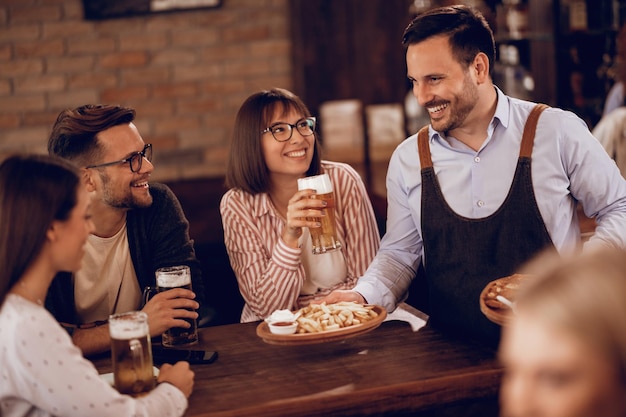 Camarero feliz sirviendo comida a un grupo de amigos mientras beben cerveza en un pub