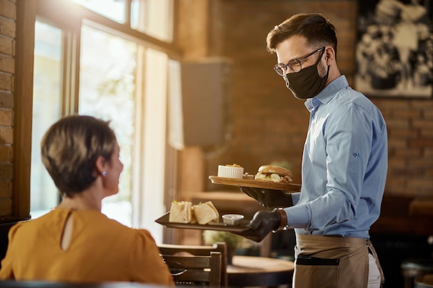 Camarero feliz sirviendo comida a un invitado mientras usa máscara protectora y guantes en un pub