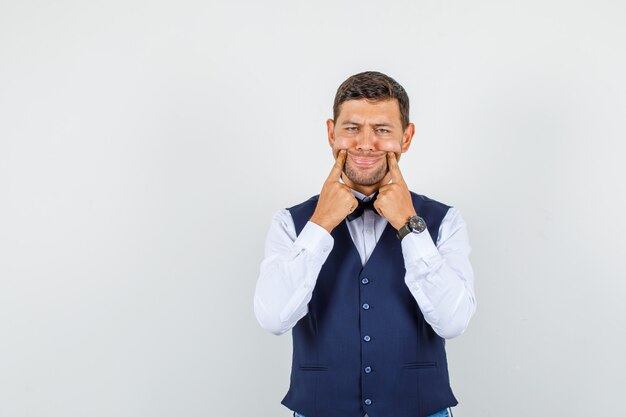 Camarero forzando una sonrisa en la cara con los dedos en camisa, chaleco y aspecto sombrío. vista frontal.