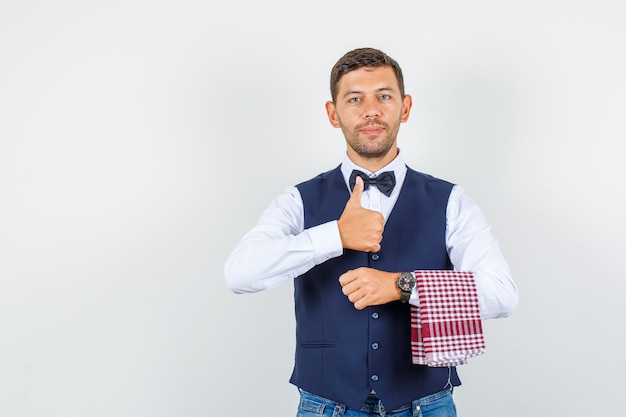 Camarero mostrando el pulgar hacia arriba en camisa, chaleco, jeans y mirando contento, vista frontal.