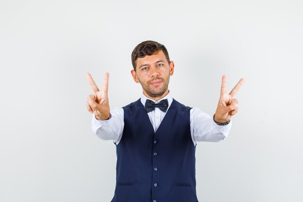 Camarero mostrando v-sign en camisa, chaleco y alegre. vista frontal.