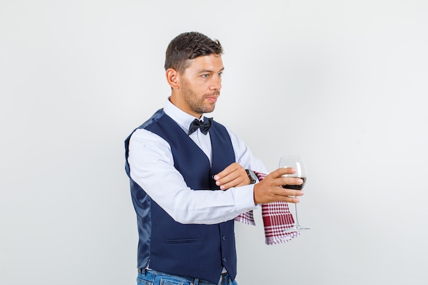 Camarero sirviendo un vaso de bebida en camisa, chaleco, jeans y mirando ocupado, vista frontal.