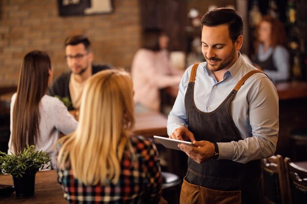 Camarero sonriente hablando con una mujer mientras toma su pedido en una tableta digital en un bar