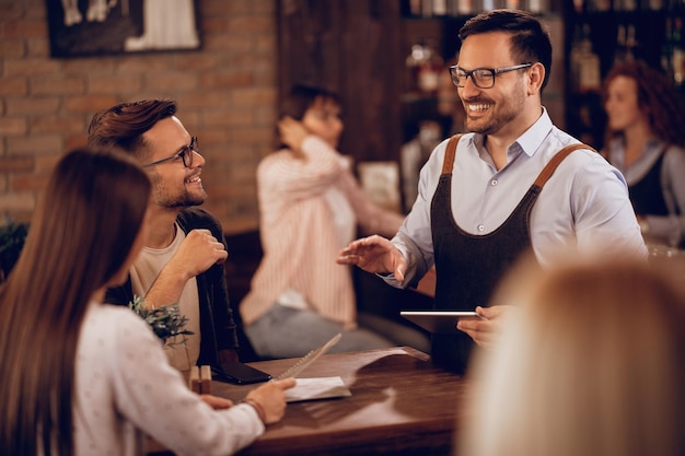 Camarero sonriente sosteniendo una tableta digital mientras toma el pedido de una pareja en un bar