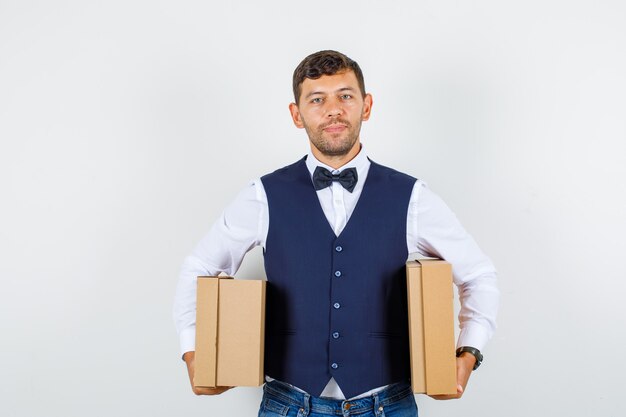 Camarero sosteniendo cajas de cartón y sonriendo en camisa, chaleco, jeans, vista frontal.