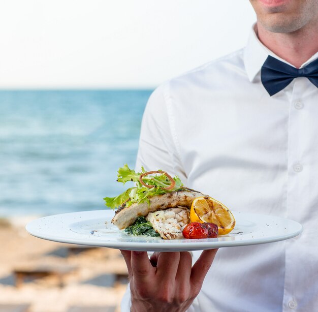 Camarero sosteniendo pescado a la parrilla con limón, tomate, hierbas cremosas en el restaurante junto al mar