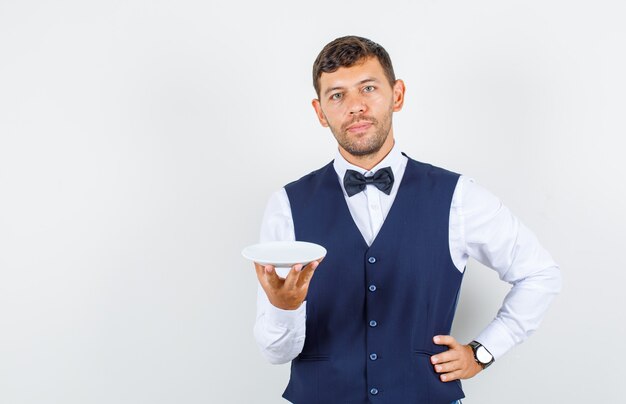 Camarero sosteniendo un plato vacío con la mano en la cintura en camisa, chaleco y mirando suave. vista frontal.