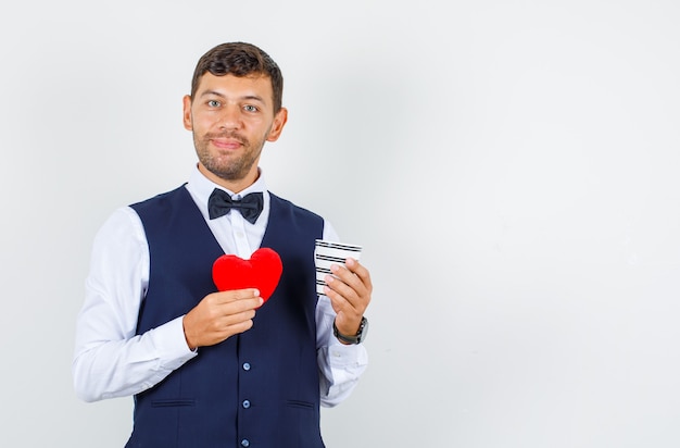 Camarero sosteniendo una taza de bebida y corazón rojo en camisa, chaleco y mirando alegre, vista frontal.