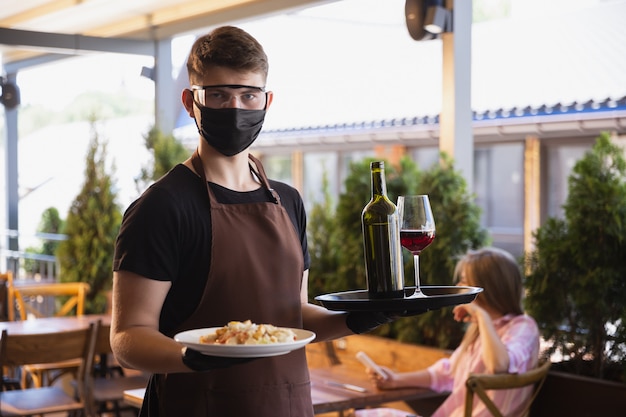 camarero trabaja en un restaurante con una máscara médica, guantes durante la pandemia de coronavirus