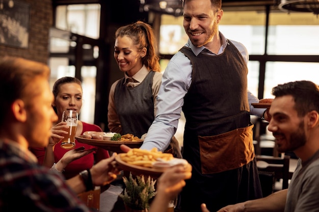 Camareros felices trayendo comida a la mesa y sirviendo a un grupo de amigos en un restaurante