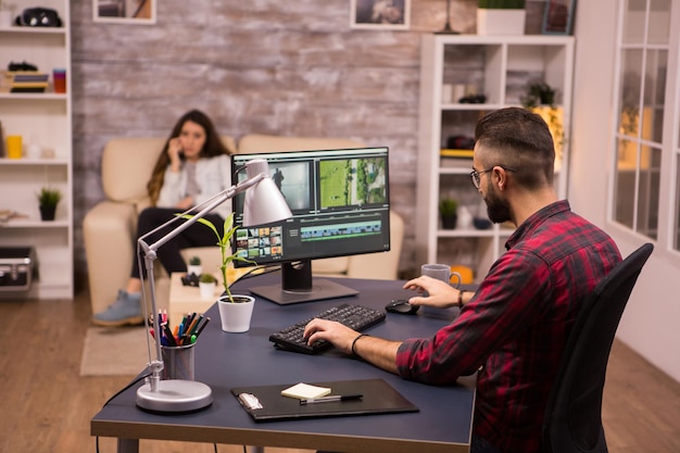 Camarógrafo creativo editando una película en la computadora desde casa. Novia en el sofá en el fondo