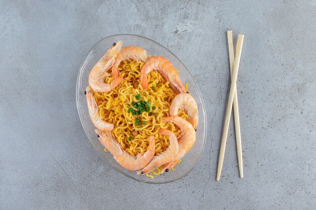 Camarones y fideos en un plato de vidrio junto a palillos, sobre la superficie de mármol.