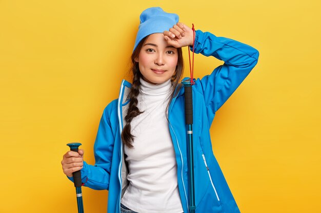 Caminante asiática cansada posa con bastones de trekking, tiene actividad al aire libre, ha viajado, vestido con traje azul, toca la frente, mira con expresión tranquila, aislado sobre una pared amarilla