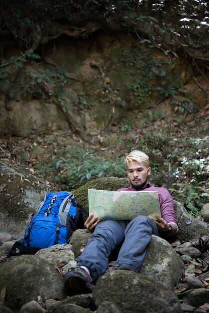 Caminante cansado joven con la mochila que se sienta en la roca en bosque mientras que se reclina después de caminata activa. Concepto de viaje