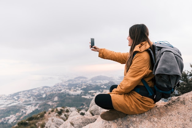 Caminante femenino joven sonriente que se sienta encima de la montaña que toma el selfie en el teléfono móvil