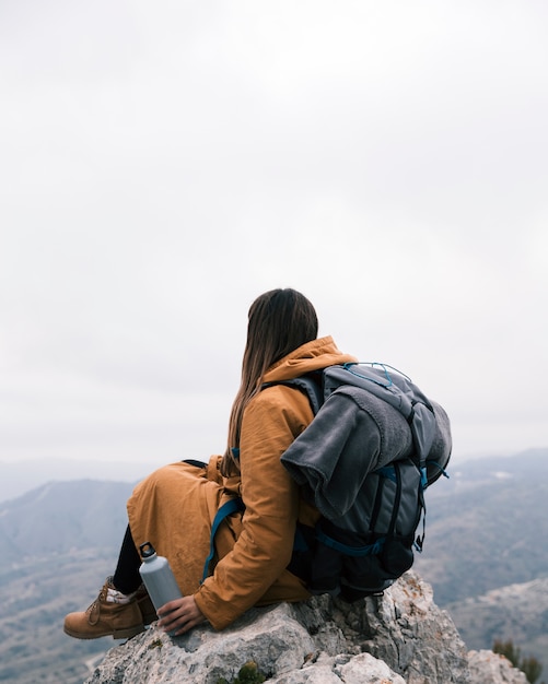 Un caminante femenino que se sienta en el pico de montaña que sostiene la botella en la mano que mira la visión
