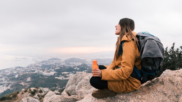 Un caminante femenino sentado en la cima de la montaña con una botella de agua en la mano con vistas a la vista