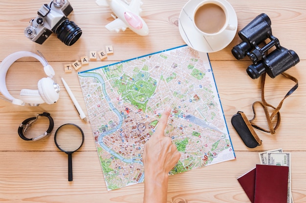 Caminante que señala en la ubicación en el mapa con una taza de té y accesorios de viajero sobre fondo de madera