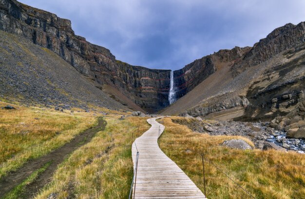 Camino de madera marrón en el campo de hierba verde cerca de la montaña rocosa marrón bajo nubes blancas durante el día