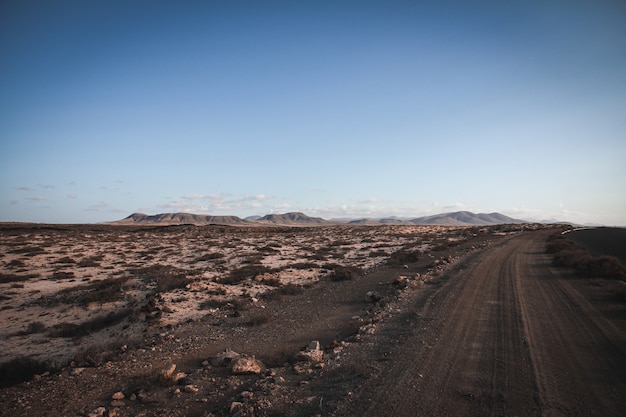 Camino de tierra cerca de un campo seco con montañas en la distancia y cielo azul claro