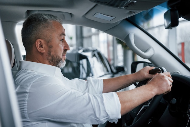 En camisa blanca y mangas enrolladas. Empresario senior en ropa oficial probando un nuevo auto de lujo en el salón del automóvil