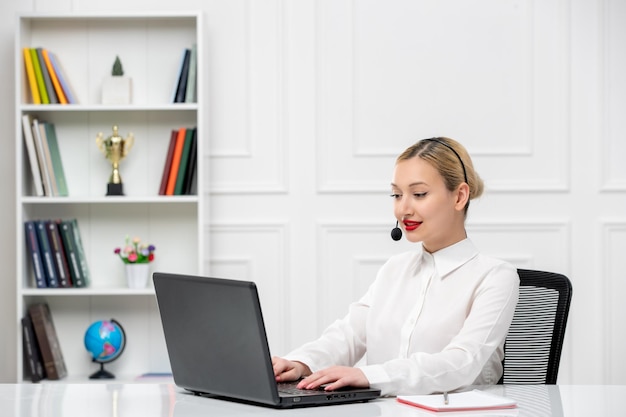 Camisa de oficina de linda chica rubia de servicio al cliente con auriculares y computadora escribiendo en la computadora portátil