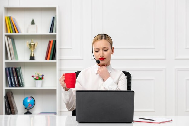 Camisa de oficina de linda chica rubia de servicio al cliente con auriculares y computadora pensando con taza roja