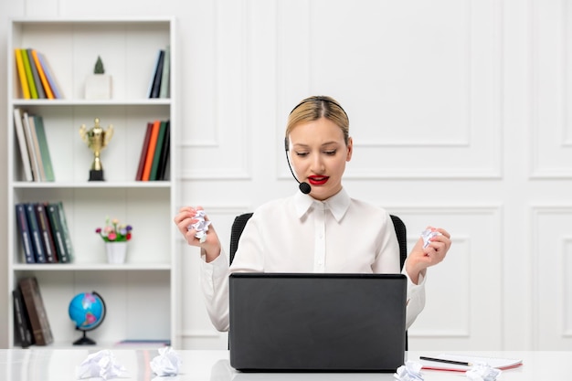 Camisa de oficina de linda chica rubia de servicio al cliente con auriculares y computadora sosteniendo un papel desmenuzado