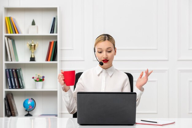 Camisa de oficina de linda chica rubia de servicio al cliente con auriculares y computadora sosteniendo una taza roja