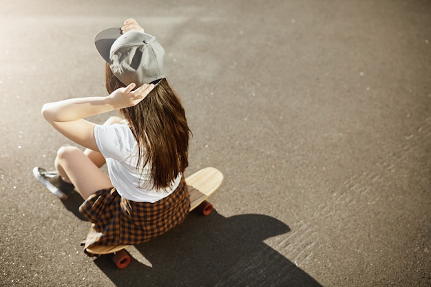 Campeona de skate femenina sentada en su longboard con sombrero en un día soleado en un entorno urbano.