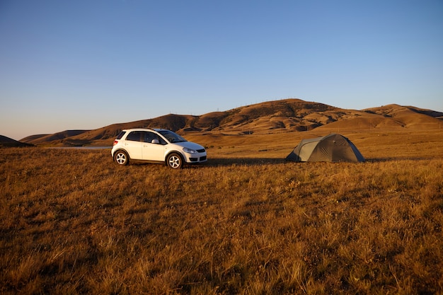 Camping en la naturaleza salvaje. Coche moderno blanco estacionado en medio del valle junto a la carpa. Turistas relajándose al aire libre, tomando un descanso durante el viaje por carretera. Hermoso paisaje de cielo azul y montañas marrones