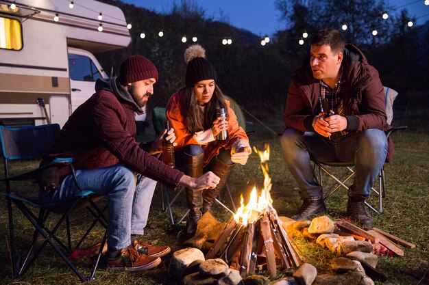 Los campistas se relajan juntos alrededor de la fogata y beben cerveza. Autocaravana retro en el fondo.