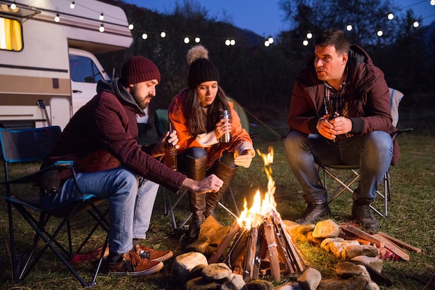 Los campistas se relajan juntos alrededor de la fogata y beben cerveza. Autocaravana retro en el fondo.