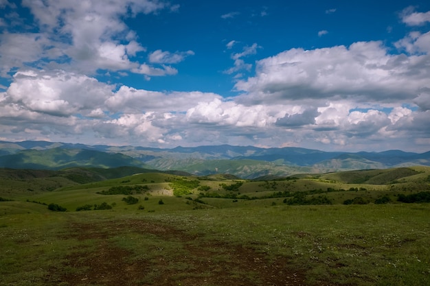 Campo cubierto de hierba rodeado de colinas cubiertas de bosques bajo un cielo nublado y la luz del sol