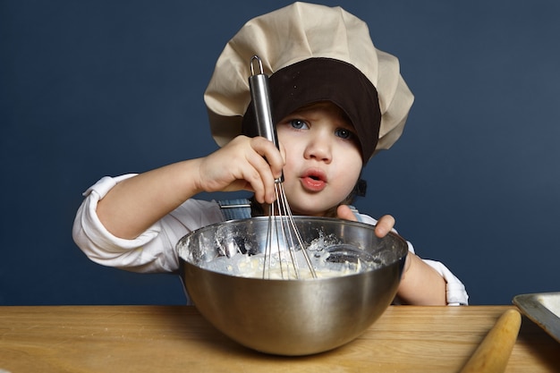 Foto gratuita cándido retrato aislado de una niña seria de 5 años en un gran sombrero de chef batiendo harina, huevos y leche en un tazón mientras hace panqueques por sí misma. receta, cocina, repostería, cocina y concepto de infancia.