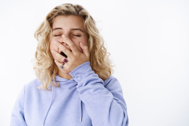 Cansada y agotada atractiva jovencita con pelo corto y rizado rubio en sudadera con capucha bostezando cubriendo la boca abierta con la palma de la mano como si quisiera tomar una siesta o dormir, de pie soñolienta sobre la pared blanca