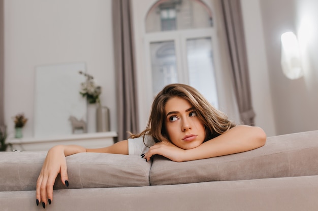 Cansada mujer de cabello castaño acostado en el sofá gris y mirando a otro lado