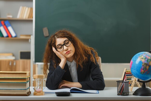 cansada con los ojos cerrados poniendo la mano en la mejilla joven maestra con gafas sentada en el escritorio con herramientas escolares en el aula