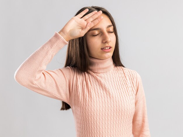 Cansado bastante adolescente manteniendo la mano en la cabeza con los ojos cerrados aislado en la pared blanca